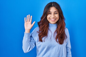 Hispanic young woman standing over blue background waiving saying hello happy and smiling, friendly...