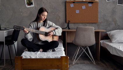 Young Asian girl plays acoustic guitar at home in her room. Learning music or musical instrument.