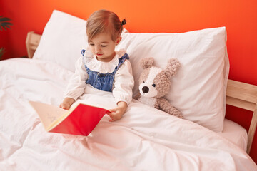 Adorable blonde toddler reading book sitting on bed with teddy bear at bedroom