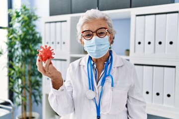 Senior grey-haired woman wearing doctor uniform and medical mask holding virus toy at clinic