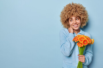 Photo of good looking woman keeps hand on neck smiles gently glad to receive flowers holds bouquet of orange gerbera dressed in casual jumper isolated over blue background blank space for your text - Powered by Adobe