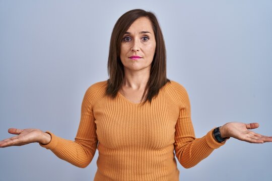 Middle age brunette woman standing wearing orange sweater clueless and confused with open arms, no idea concept.