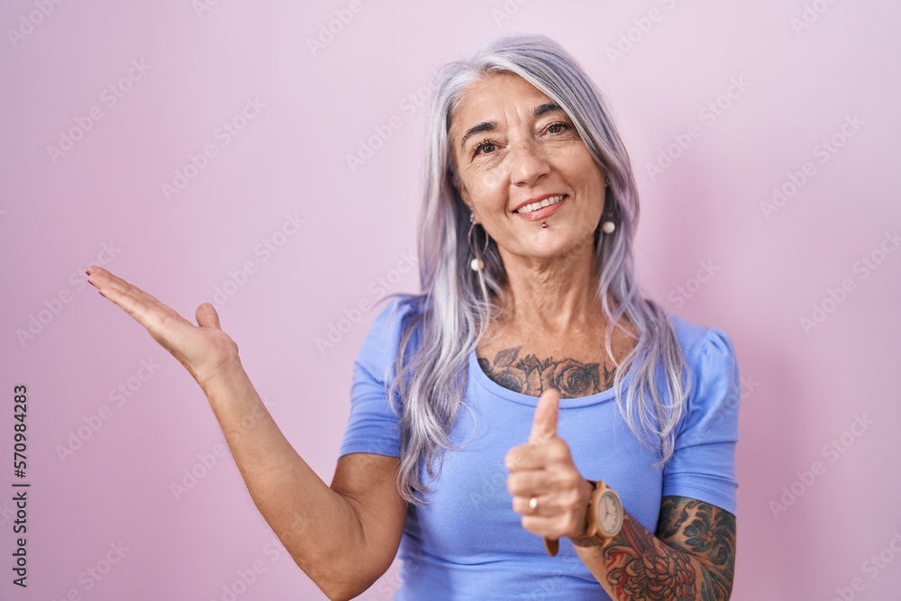 Wall mural middle age woman with tattoos standing over pink background showing palm hand and doing ok gesture w