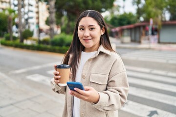 Fototapeta premium Young hispanic woman using smartphone drinking coffee at street
