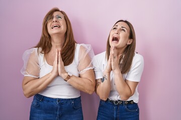 Hispanic mother and daughter wearing casual white t shirt over pink background begging and praying with hands together with hope expression on face very emotional and worried. begging.