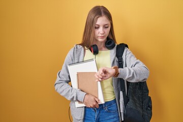 Fototapeta premium Young caucasian woman wearing student backpack and holding books checking the time on wrist watch, relaxed and confident