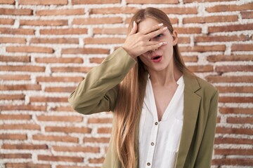 Young caucasian woman standing over bricks wall background peeking in shock covering face and eyes with hand, looking through fingers with embarrassed expression.