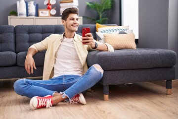 Young hispanic man using smartphone sitting on floor at home