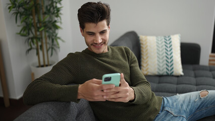 Young hispanic man using smartphone sitting on sofa at home