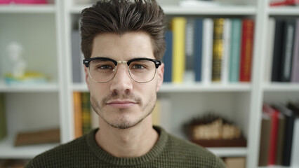 Young hispanic man student sitting on table with relaxed expression at library university