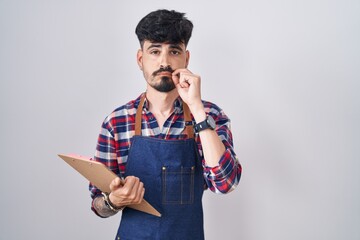 Young hispanic man with beard wearing waiter apron holding clipboard mouth and lips shut as zip...