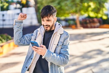 Fototapeta premium Young hispanic man using smartphone with winner expression at park