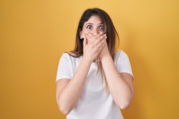 Young brunette woman standing over yellow background shocked covering mouth with hands for mistake....