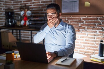 Hispanic man with beard working at the office at night shocked covering mouth with hands for...