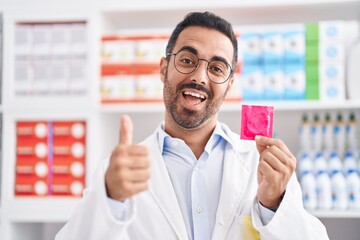 Hispanic man with beard working at pharmacy drugstore holding condom smiling happy and positive,...