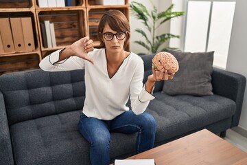 Brunette woman working at therapy office holding brain with angry face, negative sign showing...