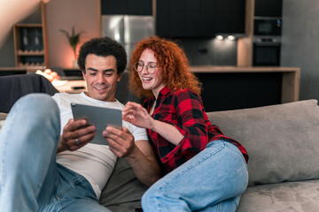 Multiracial friends are sitting very close to each other on the couch and watching funny clips on the tablet, looking affectionate