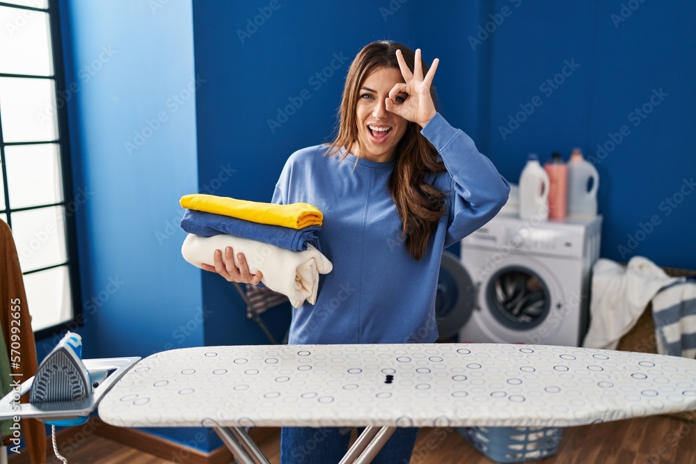 Sticker young brunette woman holding folded laundry after ironing smiling happy doing ok sign with hand on e