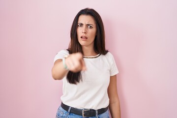 Young brunette woman standing over pink background pointing displeased and frustrated to the...