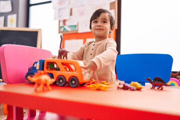 Adorable hispanic boy playing with cars and dino toys standing at kindergarten