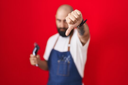 Young Hispanic Man With Beard And Tattoos Wearing Barber Apron Holding Razor Looking Unhappy And Angry Showing Rejection And Negative With Thumbs Down Gesture. Bad Expression.