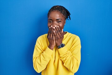 Beautiful black woman standing over blue background laughing and embarrassed giggle covering mouth...