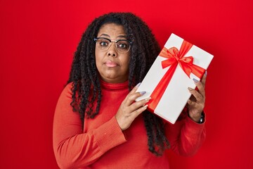 Plus size hispanic woman holding gift clueless and confused expression. doubt concept.