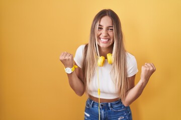 Young blonde woman standing over yellow background wearing headphones very happy and excited doing...