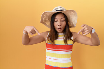 Middle age chinese woman wearing summer hat over yellow background pointing down with fingers...