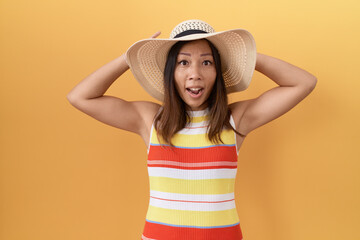 Middle age chinese woman wearing summer hat over yellow background crazy and scared with hands on...