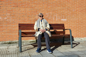 eighty year old woman in sunglasses poses sitting on a bench in front of a red brick wall