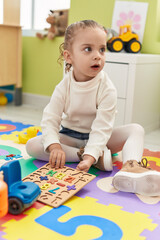 Adorable blonde girl playing with maths puzzle game sitting on floor at kindergarten