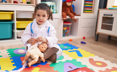 Adorable hispanic girl wearing doctor uniform bandaging arm baby doll at kindergarten