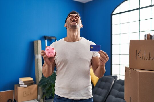 Young hispanic man with beard holding piggy bank and credit card at new home angry and mad screaming frustrated and furious, shouting with anger looking up.