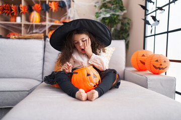 Adorable hispanic girl wearing halloween costume sitting on sofa with surprise expression at home
