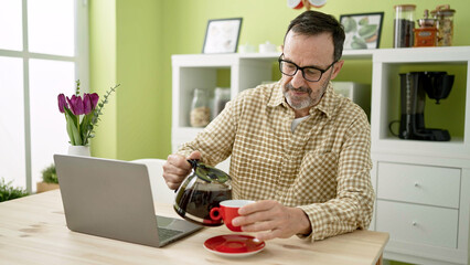 Middle age man sitting on table pouring coffee on cup at home