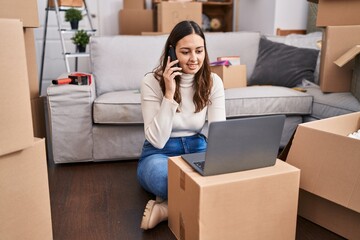 Young beautiful hispanic woman using laptop talking on smartphone at new home