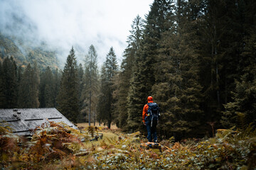 Fototapeta na wymiar A hiker with orange raincoat is exploring the surroundings of an isolated mountain hut in the forest of Val di Genova, during a rainy and foggy autumnal day, Trentino Alto Adige, Northern Italy