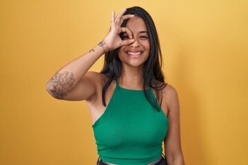 Brunette woman standing over yellow background doing ok gesture with hand smiling, eye looking through fingers with happy face.