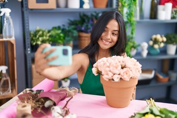 Young hispanic woman florist make selfie by smartphone sitting on table at florist shop