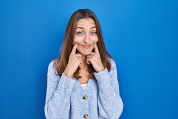 Young woman standing over blue background smiling with open mouth, fingers pointing and forcing cheerful smile