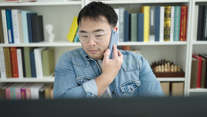 Young chinese man student using computer talking on smartphone at library university