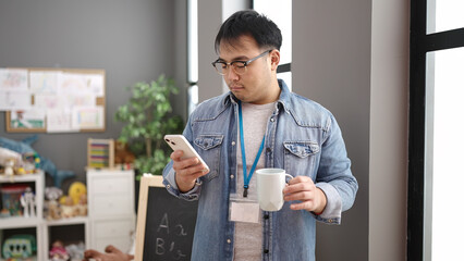 Young chinese man preschool teacher using smartphone drinking coffee at kindergarten