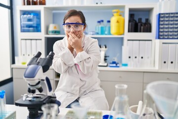 Hispanic girl with down syndrome working at scientist laboratory shocked covering mouth with hands for mistake. secret concept.