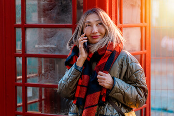 Fototapeta premium Outdoor portrait of an Asian woman visiting the UK, using a mobile phone and making a selfie near a red phone box in a city