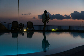 Saxophonist next a pool by sunset in tropical resort