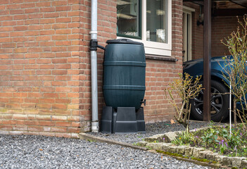 Rain barrel in front of a modern house, rainwater tank to collect rainwater and reuse it in the garden - obrazy, fototapety, plakaty