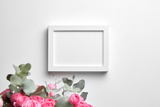 Photo frame mockup hanging on white concrete wall and bouquet of pink roses with eucalyptus leaves. Blank picture frame, interior decor