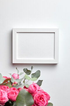 Photo frame mockup hanging on white concrete wall and bouquet of pink roses with eucalyptus leaves. Blank picture frame, interior decor