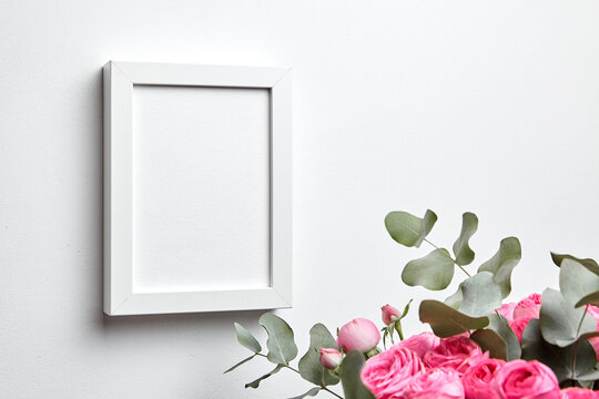 Photo frame mockup hanging on white concrete wall and bouquet of pink roses with eucalyptus leaves. Blank picture frame, interior decor